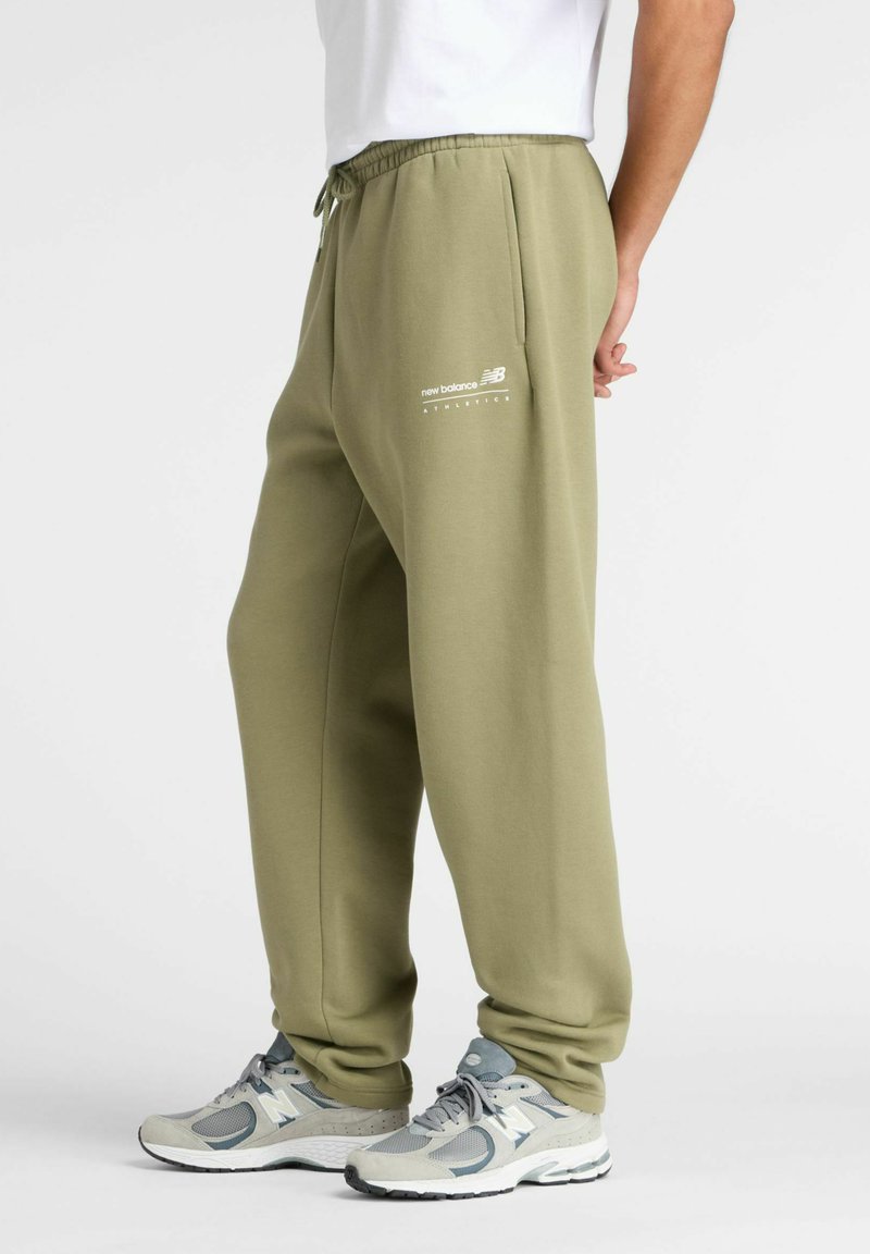 Olijfgroene sweatpants met elastische tailleband, zijzakken en wit "New Balance" logo. Gecombineerd met grijze sportschoenen.