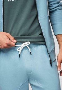 Homme ajustant le cordon blanc d'un pantalon de survêtement bleu clair, portant un sweat à capuche zippé assorti et un t-shirt vert foncé avec texte blanc partiel.