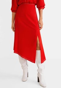 Jupe asymétrique rouge avec une fente sur le côté, en tissu lisse. Associée à des bottes noires brillantes hauteur genoux. Design minimaliste sans motifs.