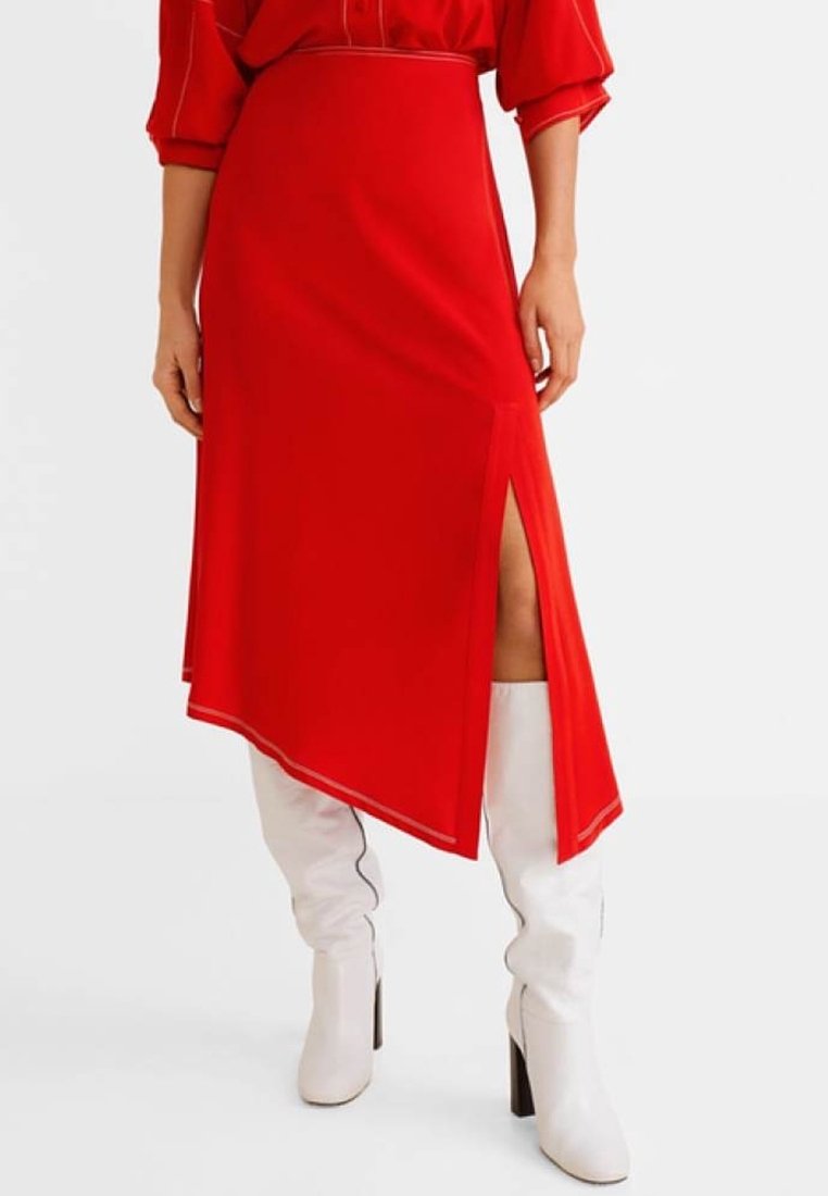 Jupe asymétrique rouge avec une fente sur le côté, en tissu lisse. Associée à des bottes noires brillantes hauteur genoux. Design minimaliste sans motifs.