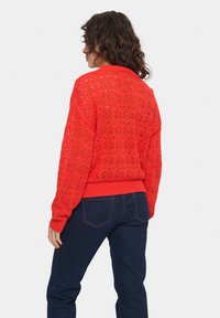 Maglione rosso a maglia con motivo di pizzo aperto, orlo e polsini a coste, indossato con jeans di denim scuro. La vista posteriore mostra un intricato design della texture.