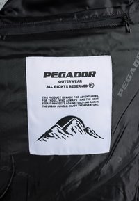 Materialul exterior negru cu un eticheta albă care prezintă logo-ul "PEGADOR", instrucțiuni de întreținere și un grafic cu munți. Textul subliniază pregătirea pentru aventuri.
