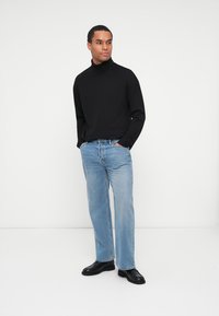 Maglione nero a maniche lunghe con colletto a dolcevita abbinato a jeans dritti blu chiaro e scarpe nere, il tutto su uno sfondo neutro.