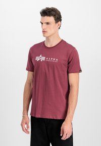 Tumši sarkans T-krekls ar balto "ALPHA INDUSTRIES" logotipu, apaļu kakla izgriezumu, īsām piedurknēm un mazu birku uz kreisās piedurknes.