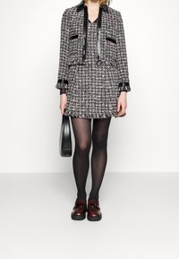 Růžový a černý tvídový šaty s odpovídajícím cropped sakem, které má třásně a černé kožené detaily. Modelka má na sobě černé punčocháče a burgundzské boty.