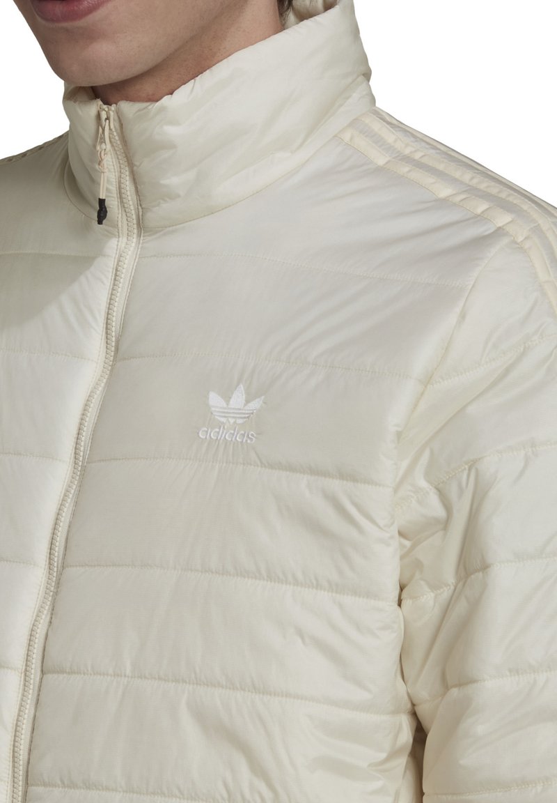 Doudoune beige clair avec motif matelassé, col montant, fermeture éclair intégrale et logo Adidas blanc discret sur la poitrine.