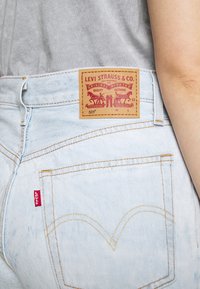 Vaqueros Levi's 501 de color azul claro con costuras visibles en los bolsillos traseros y parche de cuero de la marca, usados con una camisa gris y el brazo al lado de la cintura.