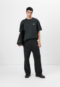 Svart oversized t-skjorte med en liten neongrønn logo, kombinert med løse svarte bukser og en svart totebag. Glatt stoff, minimalistisk design.