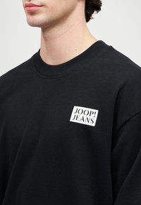 Felpa nera realizzata in tessuto morbido, con un colletto a coste e un logo rettangolare bianco con "JOOP! JEANS" stampato in nero.