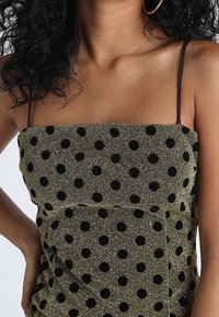 Svart prickig möster på ett skimrande olivgrönt tyg; figursydd design med tunna axelremmar och rak halsringning.