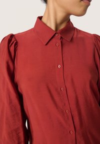 Red shirt with a classic collar, button-down front, and puffed sleeves. Made of smooth fabric with a soft sheen and red buttons.