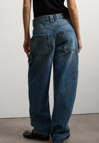 Persona vestida con jeans de mezclilla azul holgados, con grandes bolsillos traseros y mocasines negros, de pie contra una pared blanca sencilla.