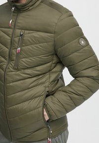 Gepolsterte Jacke in Olivgrün mit diagonalen Steppmustern, hohem Kragen, zwei seitlichen Reißverschlusstaschen und Logo-Patch am Ärmel.