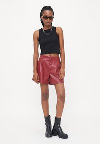 Top senza maniche nero, pantaloni in pelle rossi con un design plissettato e stivaletti neri alla caviglia. La modella è accessoriata con occhiali da sole e una collana a catena.