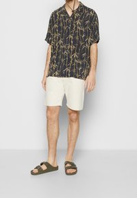 Camicia a maniche corte blu navy con motivo giallo a bambù, abbinata a pantaloni corti crema e sandali scivolosi verde scuro. Chiusura semplice con bottone al colletto.