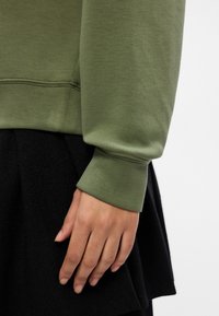 Sudadera verde oliva con un puño texturizado, combinada con una prenda negra. La tela parece suave, con un ajuste ceñido alrededor de la muñeca.