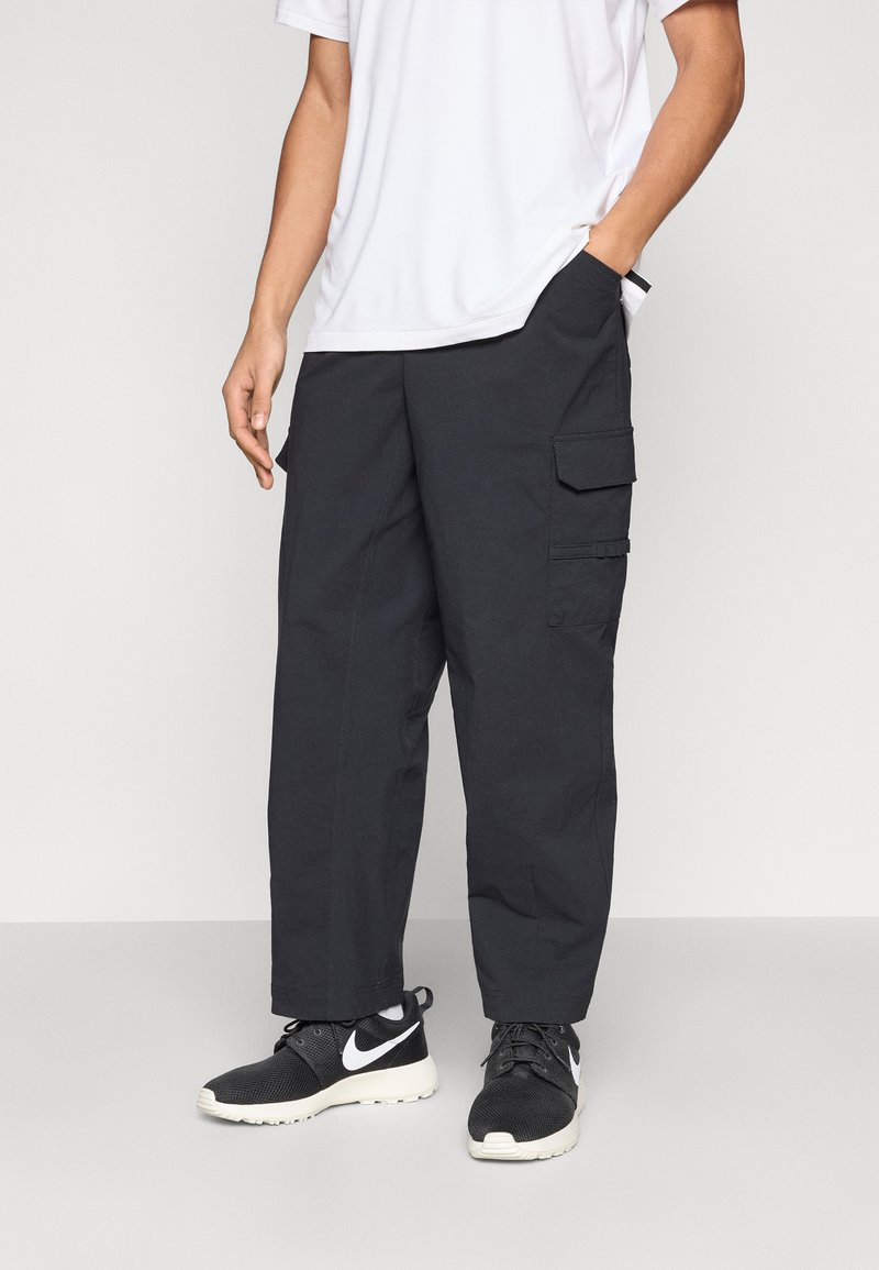 Fekete cargo nadrág több oldalzsebbel, tartós anyagból készült. Fehér pólóval és fekete sportcipővel párosítva.