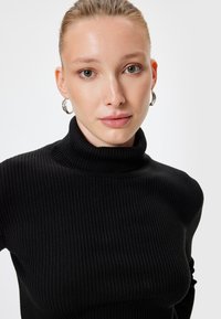 Maglione aderente a collo alto nero a coste. Il tessuto appare texturizzato con righe verticali e un colletto alto. Sono visibili orecchini a cerchio di argento.