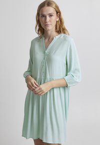 Rochie verde mentă deschisă, confecționată dintr-un material neted, cu un decolteu cu nasturi, guler în formă de V, mâneci 3/4 și o talie plisată pentru o croială lejeră.