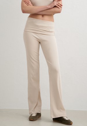 Persona con pantalones beige de cintura alta y pierna ancha y top corto beige con los brazos cruzados, de pie sobre un suelo gris frente a una pared blanca.
