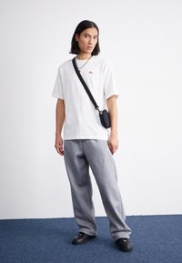 T-shirt bianco oversize con piccolo logo rosso, pantaloni della tuta grigi con una vestibilità ampia, borsa a tracolla nera e scarpe nere slip-on.