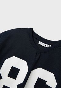 Granatowa siatkowa koszulka sportowa z białym nadrukiem "86" z przodu. Posiada ściągacz przy okrągłym dekolcie oraz teksturowany materiał. Widoczna metka marki.