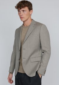 Brun och vit rutig blazer med slag, två framfickor och knäppta manschetter, bärs över en beige rundhalsad tröja.