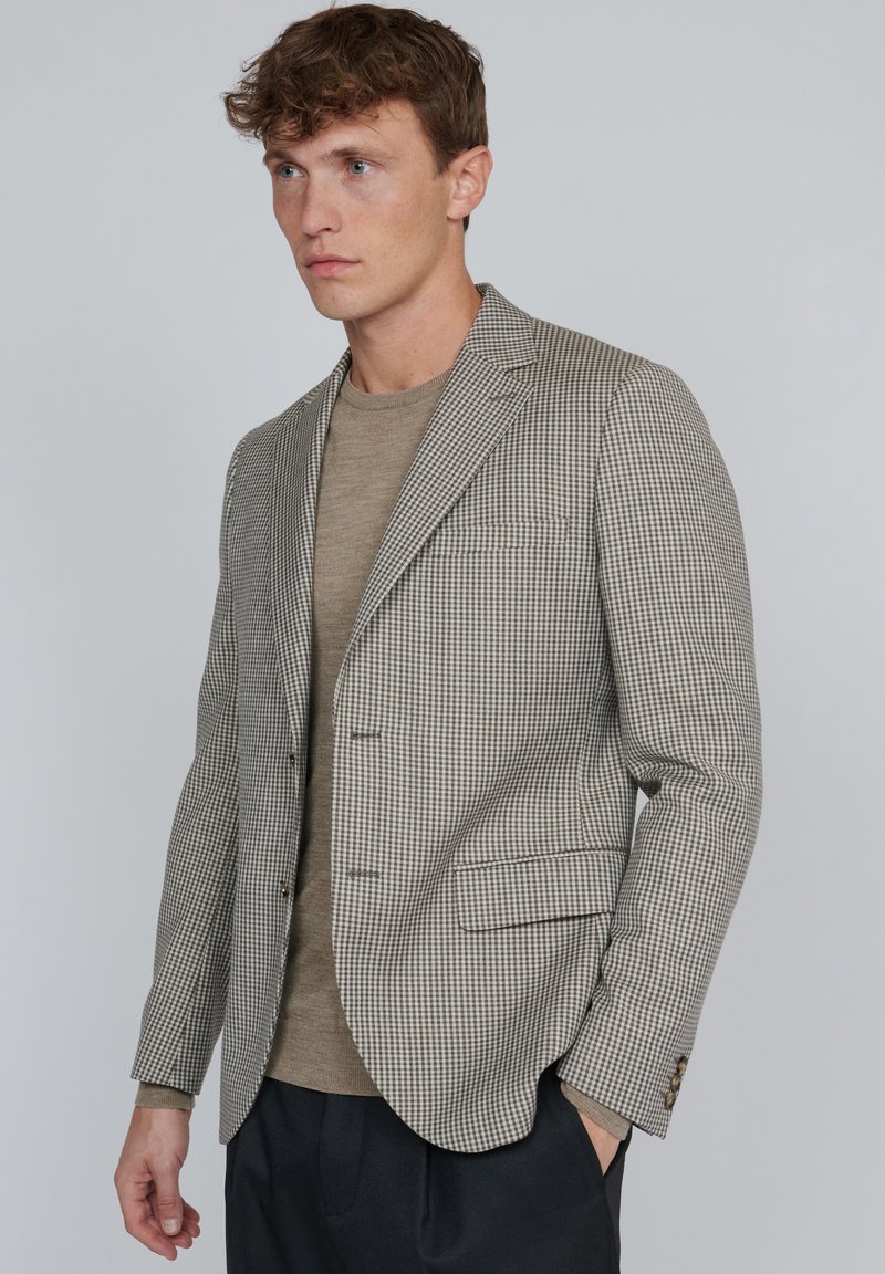 Brun och vit rutig blazer med slag, två framfickor och knäppta manschetter, bärs över en beige rundhalsad tröja.