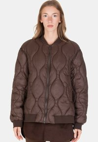 Donna che indossa una giacca trapuntata marrone con zip, polsini e orlo a coste, in piedi davanti a uno sfondo chiaro e semplice.