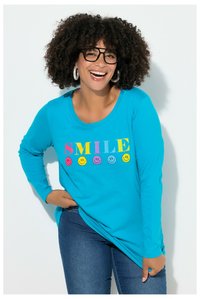 Lächelnde Frau mit lockigem Haar und Brille, die blaue Jeans und ein hellblaues Langarmshirt mit buntem "SMILE"-Schriftzug und Smiley-Gesichtern trägt.