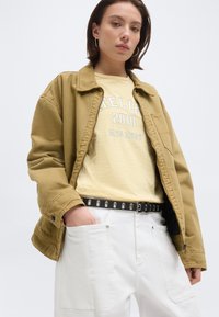 Νέα γυναίκα που φοράει μπεζ μπουφάν, κίτρινο ριγέ πουκάμισο με κείμενο, λευκό παντελόνι και μαύρη ζώνη, με τα χέρια εν μέρει στις τσέπες, ουδέτερο φόντο.