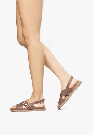 Sandales marron avec boucle réglable, dotées de larges brides croisées et d'une semelle plate. Texture lisse, design minimaliste et bout ouvert.