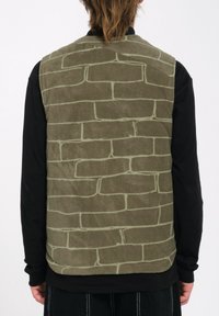 Chaleco sin mangas verde con un patrón texturizado de ladrillos en la espalda, hecho de tela suave, llevado sobre una camiseta negra de manga larga.