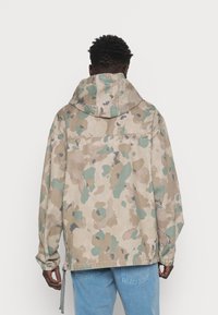 Camo-mønstrede, oversized jakke i beige, grønne og blå nuancer. Har hætte, elastiske manchetter og snoretræk i kanten.
