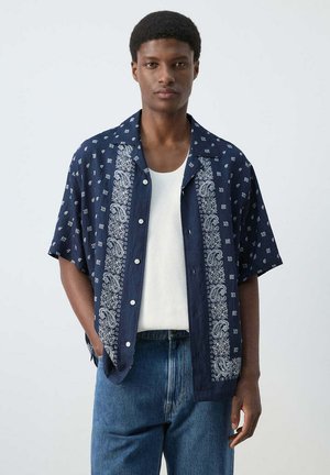 Joven con camisa azul marino de manga corta y botones, con patrones blancos de cachemira, sobre camiseta blanca sin mangas y vaqueros azules.