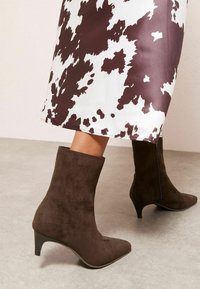 Rudos zomšinės aulinės batai su smailiu priekiu ir 5 cm kūgio formos kulnu, derinami su raštuota balta ir ruda suknele.