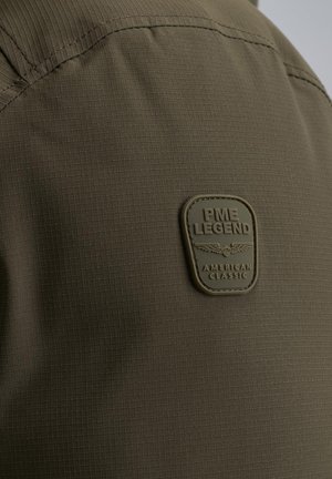Tela de color verde oliva con un patrón de cuadrícula texturizada, que presenta un parche rectangular de goma con texto en relieve: "PME LEGEND AMERICAN CLASSIC."