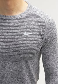 Grijze sporttop met een gestructureerd patroon over de borst, ronde halslijn en witte Nike-logo aan de linkerkant.