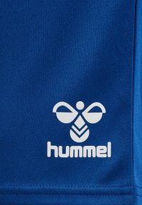 White stylized bee logo and "hummel" text on royal blue textured fabric background, likely sportswear material.