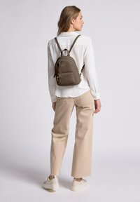 Mochila de cuero marrón con dos bolsillos con cremallera, correas ajustables y una forma redondeada. El modelo lleva una camisa crema y pantalones beiges.