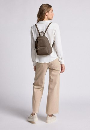Brauner Leder-Rucksack mit zwei Reißverschlussfächern, verstellbaren Trägern und runder Form. Das Modell trägt ein cremefarbenes Shirt und beige Hosen.
