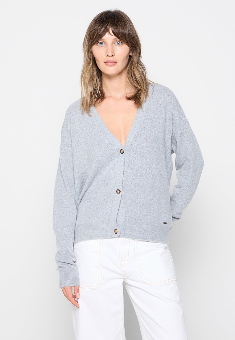 Cardigan grigio chiaro, oversize, realizzato in materiale morbido, con scollo a V profondo, maniche lunghe e grandi bottoni frontali.