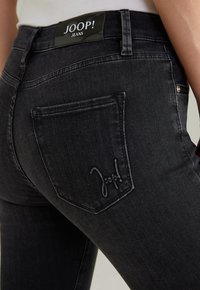 Jean en denim foncé avec une coupe ajustée, une taille haute, un logo brodé sur la poche arrière et une texture lisse.