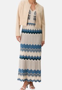 Chaqueta de ante sintético beige sobre un vestido largo estampado con diseño de zigzag en beige, azul y verde azulado, con corpiño ajustado y falda fluida.