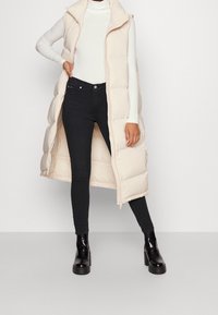 Chaleco sin mangas largo y acolchado en color beige claro con cremallera frontal y cuello alto, combinado con jeans ajustados negros y botas negras gruesas.