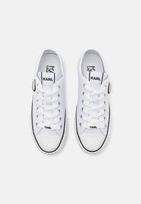Un paio di sneakers bianche stringate con rifiniture nere e il marchio "KARL" sulle linguette e sui lacci, viste dall'alto su uno sfondo neutro.