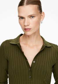 Young woman with slicked-back hair wearing a ribbed olive green buttoned shirt and a silver hoop earring, looking at the camera.