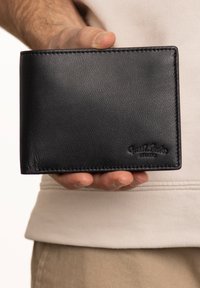Black leather wallet held in a hand, featuring a smooth texture, straight edges, and embossed branding on the lower right corner.