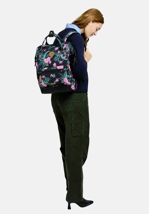 Young woman wearing dark cargo pants, navy sweater, and floral backpack, looking down against a plain light background.