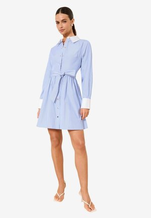 Frau, die in einem blau gestreiften Hemdblusenkleid mit weißem Kragen und Manschetten, gebundener Taille und weißen Sandalen mit Absatz vor weißem Hintergrund steht.